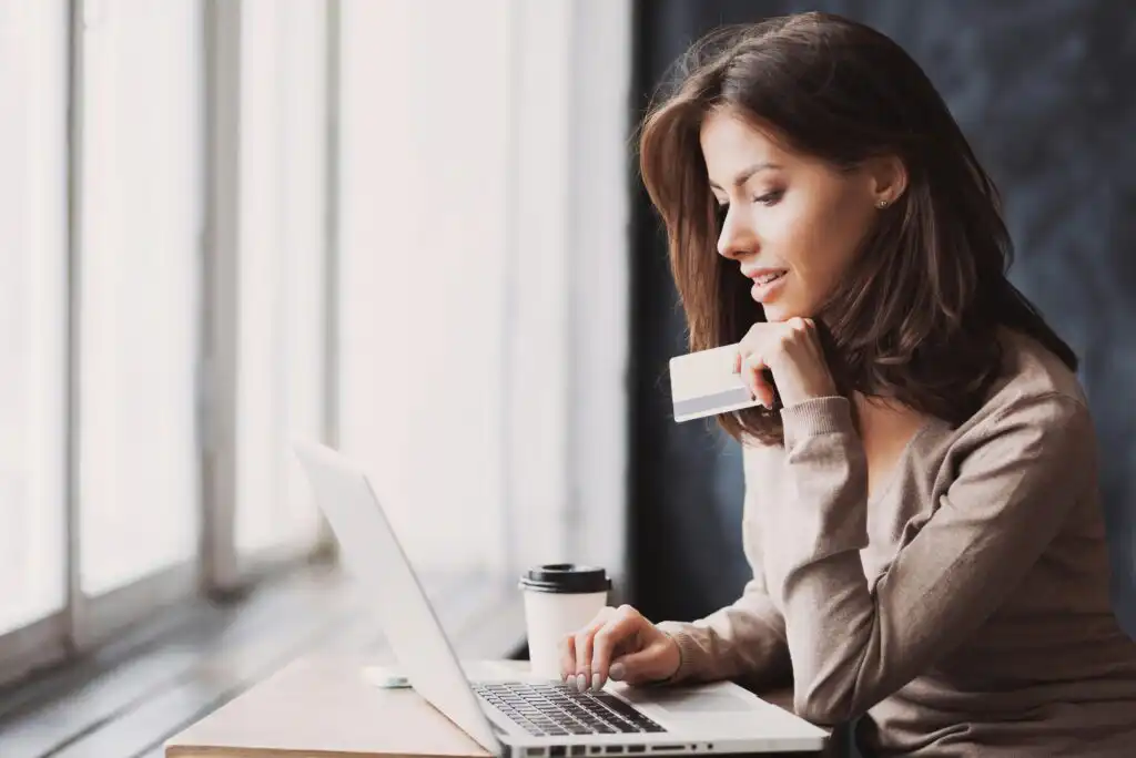 A woman sits at a table by a window, holding a credit card and using a laptop. A coffee cup is on the table beside her. She appears to be smiling and focused on the screen.