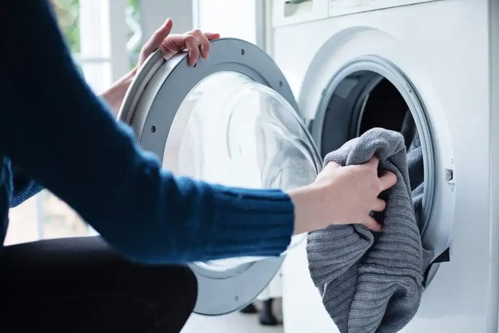 A person in a blue sweater loads a gray sweater into a front-loading washing machine, with the washing machine door open.
