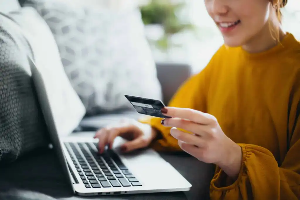 A person wearing a yellow top is sitting on a couch, smiling while holding a credit card in one hand and using the other hand to type on a laptop.