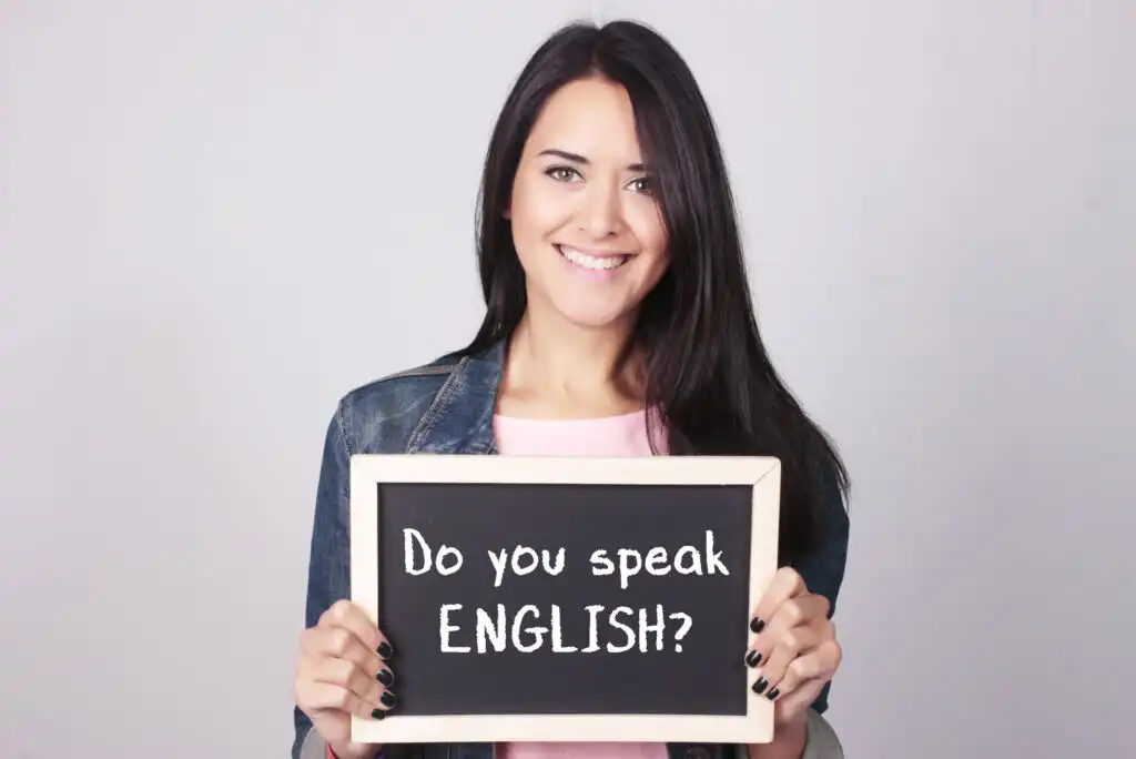 A woman with long dark hair smiles and holds a chalkboard sign that reads, Do you speak ENGLISH? against a plain light background.