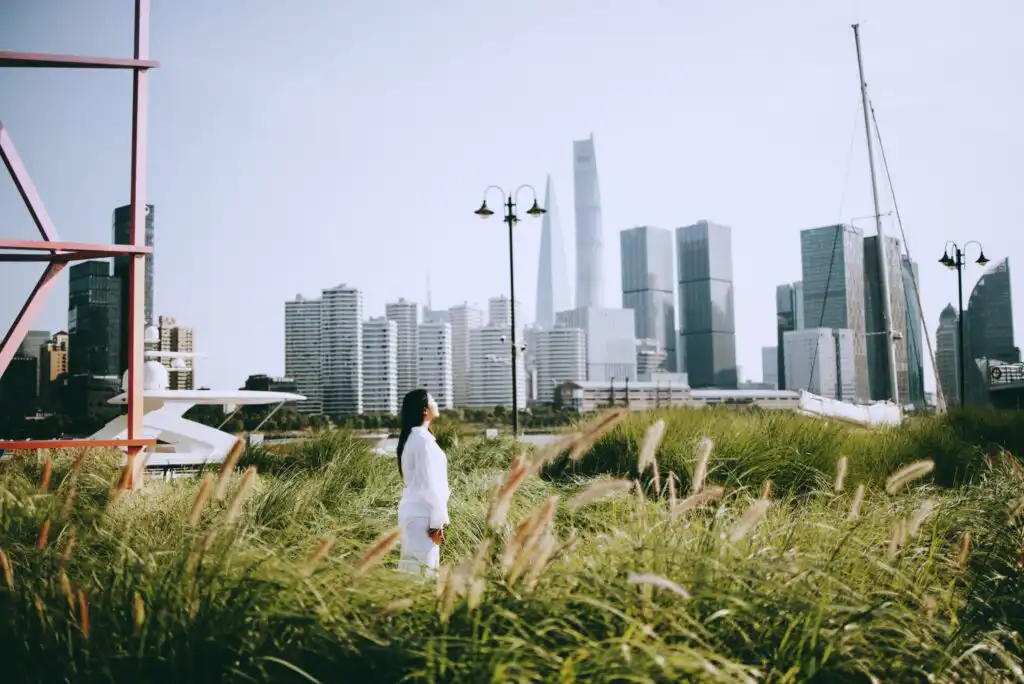 A woman in a white outfit stands in tall grass, gazing at modern city skyscrapers in the background on a clear day. A yacht, lamp post, and sailboat mast are also visible, blending nature with an urban skyline.