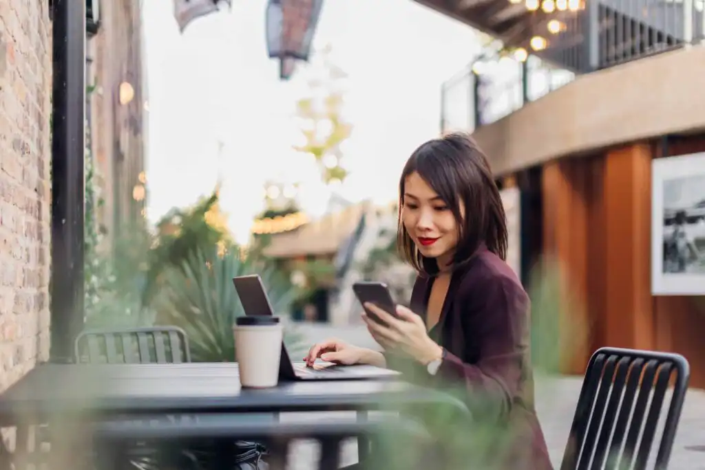 A woman sits at an outdoor table with a laptop, holding a smartphone and smiling. A coffee cup is on the table, and the background features plants and string lights in a modern outdoor setting.