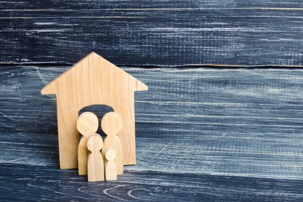 Wooden figures representing a family of four stand inside a wooden house shape, set against a dark wooden background with horizontal planks.