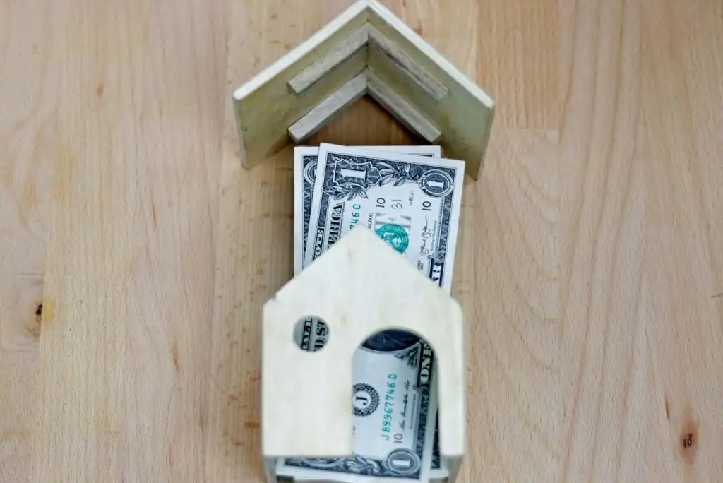 A small wooden house frame sits on a wooden surface, surrounding several one dollar bills, symbolizing real estate investment or home financing.