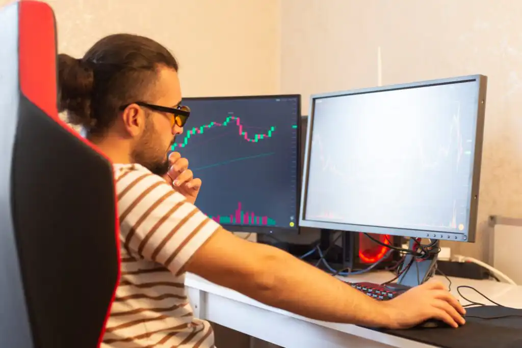 A man wearing glasses and a striped shirt sits at a desk with two computer monitors, analyzing stock charts and financial graphs on the screens.