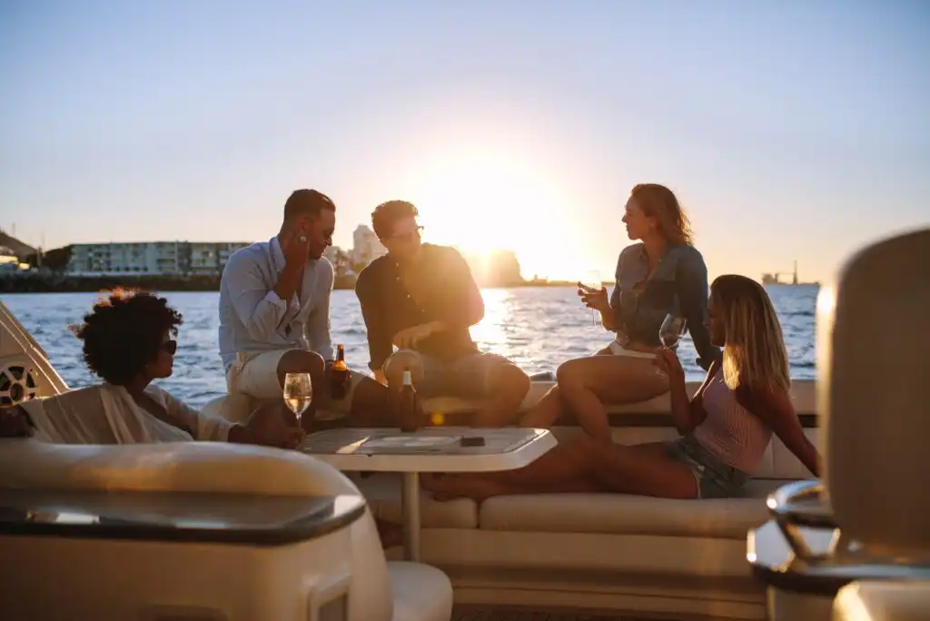 Five people relax and socialize on a yacht at sunset, enjoying drinks and conversation with a scenic waterfront and city buildings in the background. The atmosphere is warm and leisurely.