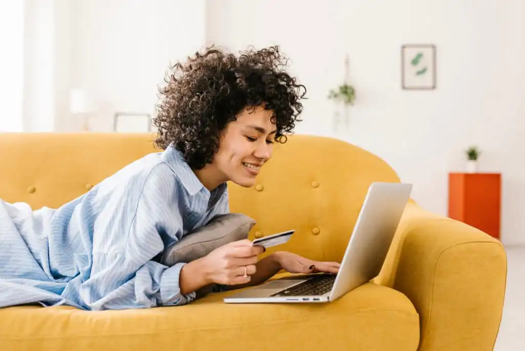 A person lying on a yellow couch smiles while holding a credit card and using a laptop, suggesting online shopping or banking in a bright, cozy living room.