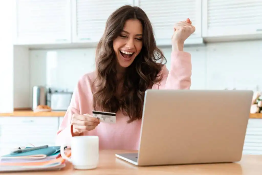 A woman sitting at a table in a kitchen smiles excitedly at her laptop, holding a credit card in one hand and raising her other fist in celebration. A mug, notebooks, and pen are on the table.