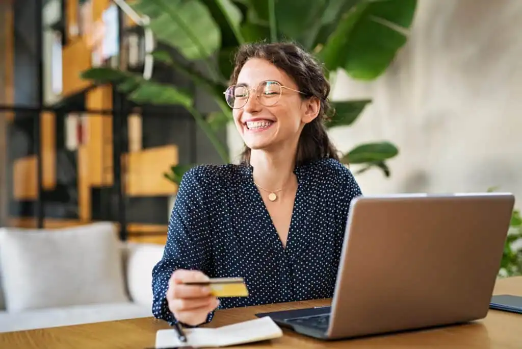 A smiling woman with glasses sits at a desk with a laptop, holding a credit card in one hand and a notebook in front of her. There are large green plants and shelves in the background.