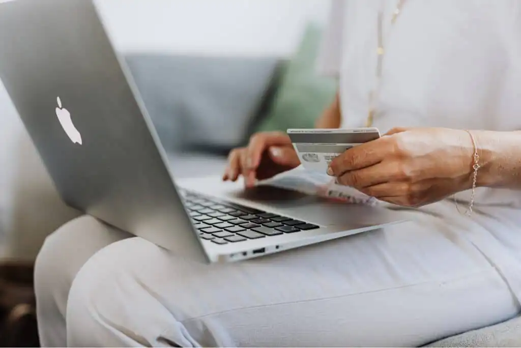 A person sitting with a laptop on their lap holds a credit card in one hand and types on the keyboard with the other, suggesting online shopping or payment.
