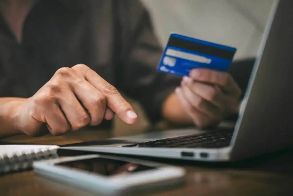 A person holds a credit card in one hand and uses the other hand to type on a laptop keyboard. A smartphone and a notebook are on the table nearby.