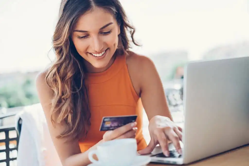 A smiling woman in an orange sleeveless top is sitting at a table with a laptop, holding a credit card and typing. A white coffee cup sits in front of her. The background is blurred, suggesting an outdoor setting.