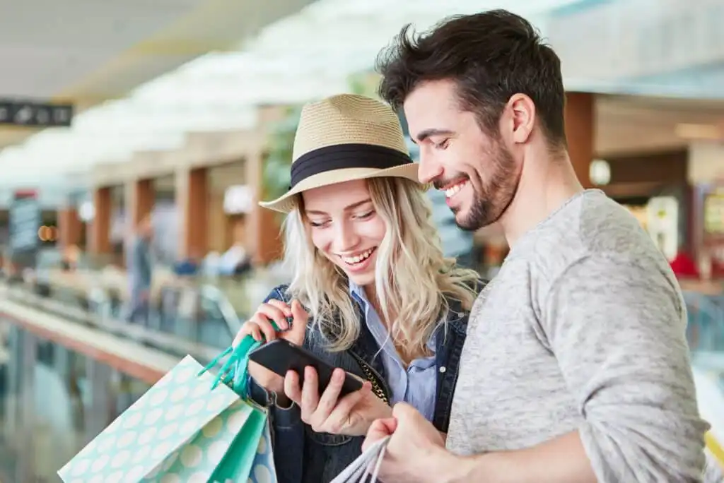 A smiling woman and man stand close together in a shopping mall, holding shopping bags. The woman wears a hat and looks at a smartphone with the man, both appearing happy and engaged.
