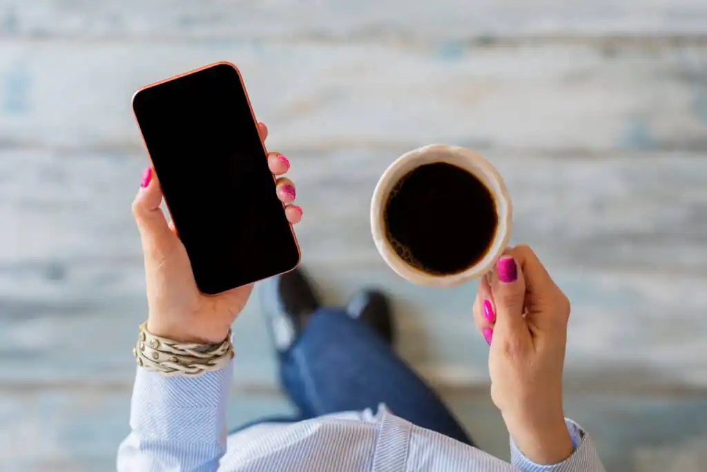 A person with pink nail polish holds a smartphone with a blank screen in one hand and a cup of black coffee in the other, standing on a light wooden floor.