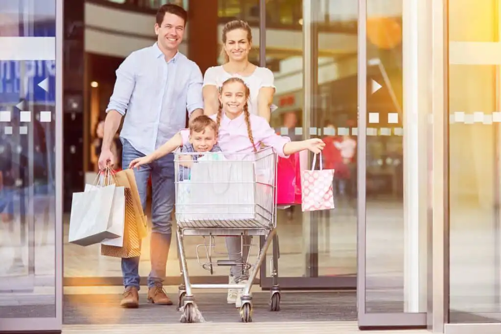 A smiling family of four exits a shopping mall. The parents carry shopping bags, while their two children ride in a shopping cart, looking happy. Sunlight streams through the glass doors behind them.