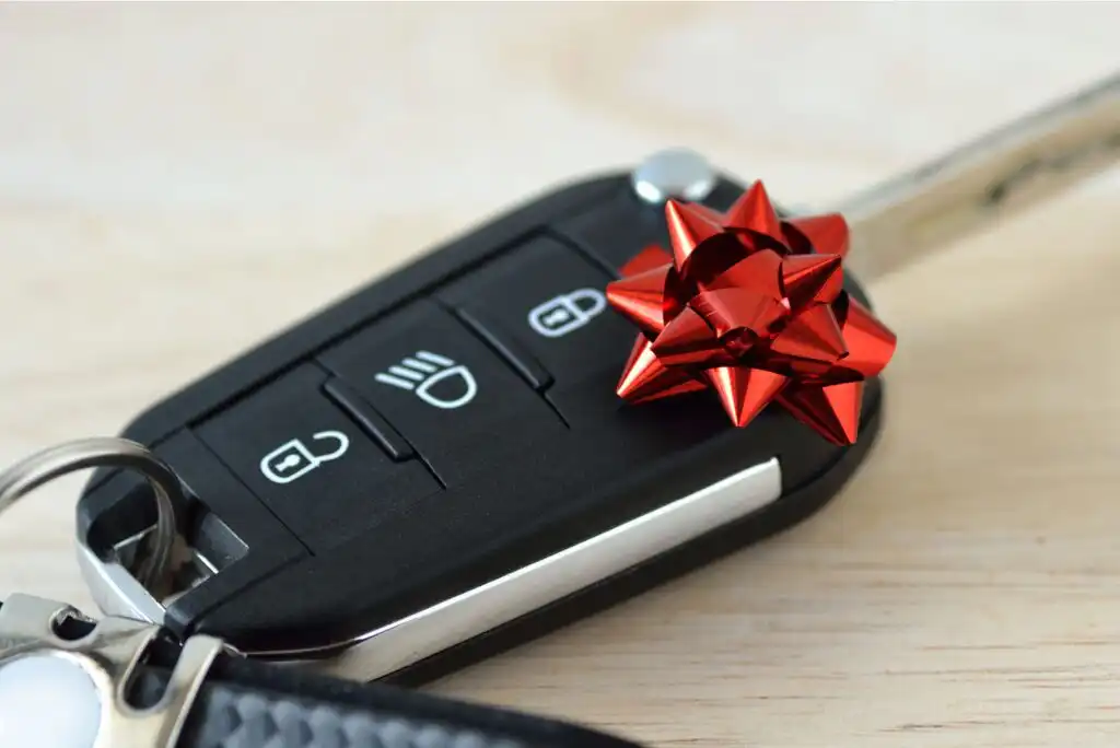 A car key with buttons for lock, unlock, and lights is decorated with a small red gift bow, resting on a light wooden surface.