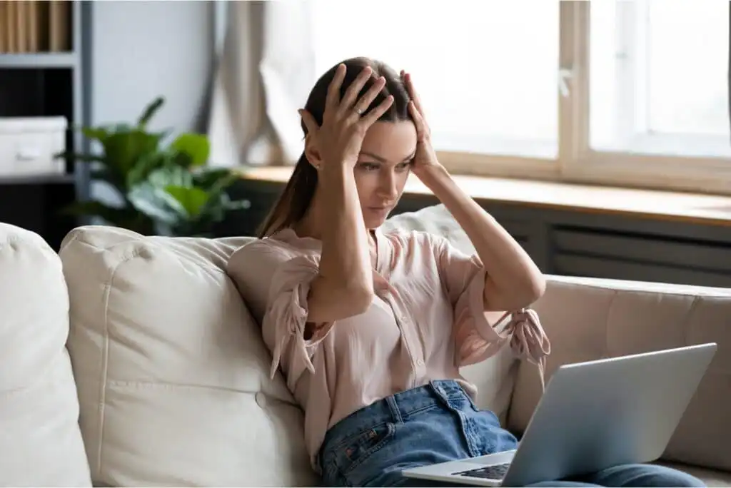 A woman sits on a couch with her hands on her head, looking stressed or frustrated while staring at a laptop screen. She is wearing a light pink blouse and jeans. A plant is visible in the background.