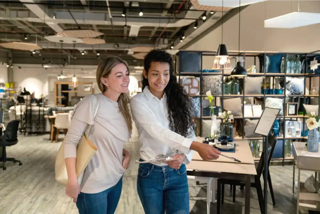 Two women smiling and looking at something off-camera in a modern home goods store. One woman holds a clipboard and gestures, while the other stands beside her with a handbag, suggesting a shopping or consultation experience.