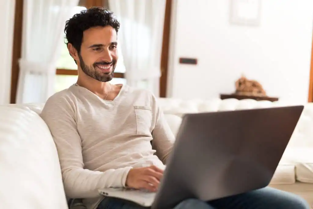 A man with curly hair and a beard sits on a white sofa, smiling while using a laptop. He is casually dressed in a light-colored shirt and jeans, with a bright, cozy room in the background.