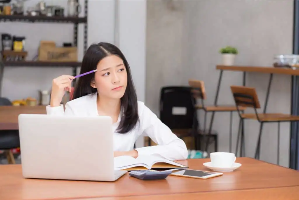 A woman sits at a table with a laptop, open book, and cup of coffee, holding a pen to her temple and looking thoughtful. Shelves and chairs are visible in the background.