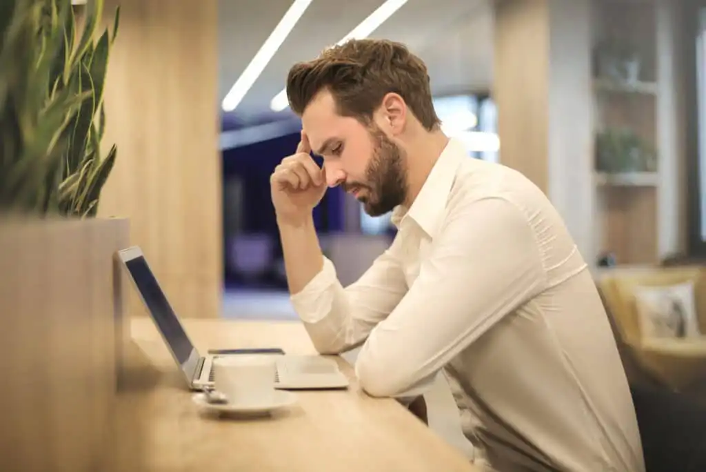 A man in a white shirt sits at a desk in front of a laptop, resting his head on his hand and looking thoughtful or stressed. A cup and saucer sit beside him, and plants are in the foreground.