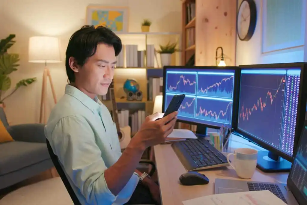 A man sits at a desk with multiple monitors displaying financial charts and graphs, holding a smartphone and smiling. The workspace is organized, with a keyboard, documents, and a coffee mug nearby.