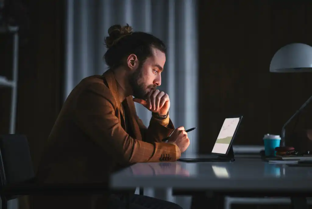 A man with a beard and tied-back hair sits at a desk, thoughtfully looking at a tablet with a stylus in hand. He wears a brown jacket in a dimly lit room with a coffee cup on the table.