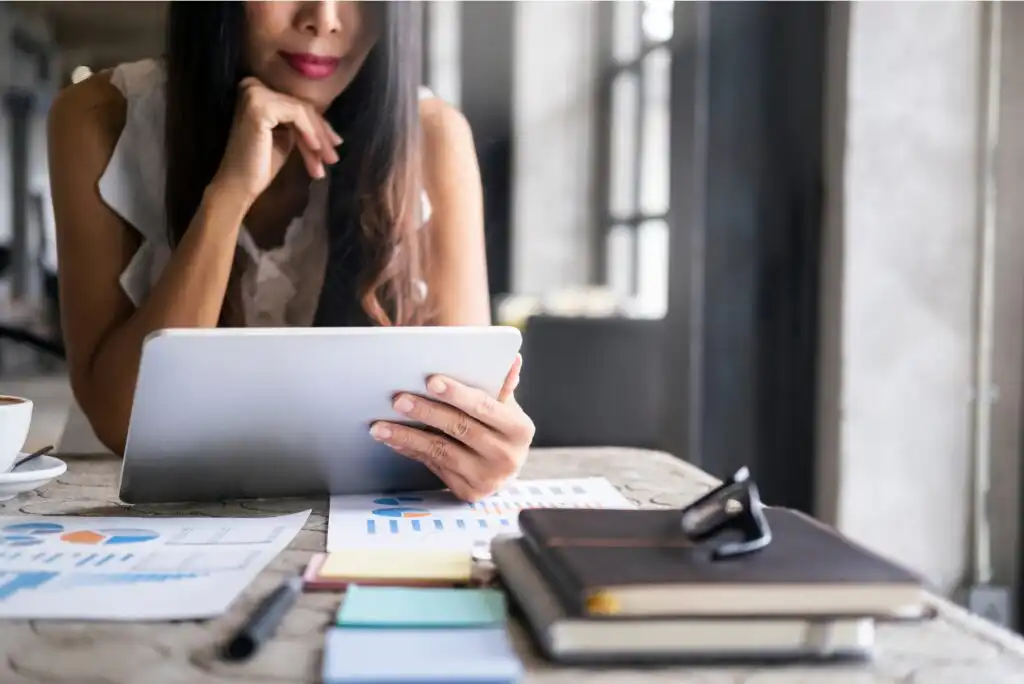 A woman sits at a table using a tablet, with business charts, notebooks, and sticky notes spread in front of her. She appears focused, resting her chin on her hand in a well-lit, modern workspace.