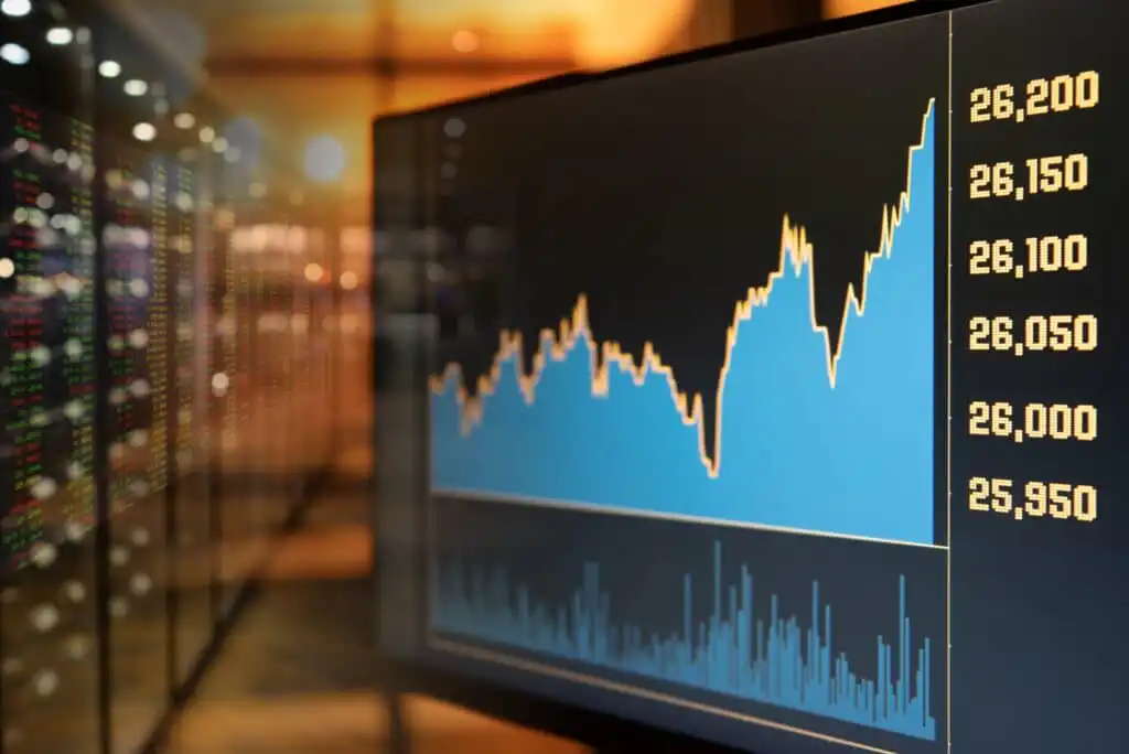 A close-up of a computer screen displaying a rising stock market graph with numerical values on the right, reflecting financial growth. Blurred server racks or screens are visible in the background.