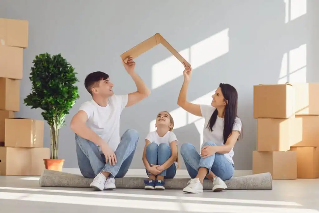 A smiling family of three sits on the floor among moving boxes, holding a cardboard roof shape over their heads, symbolizing a new home. All wear white shirts and jeans. A green potted plant is in the background.