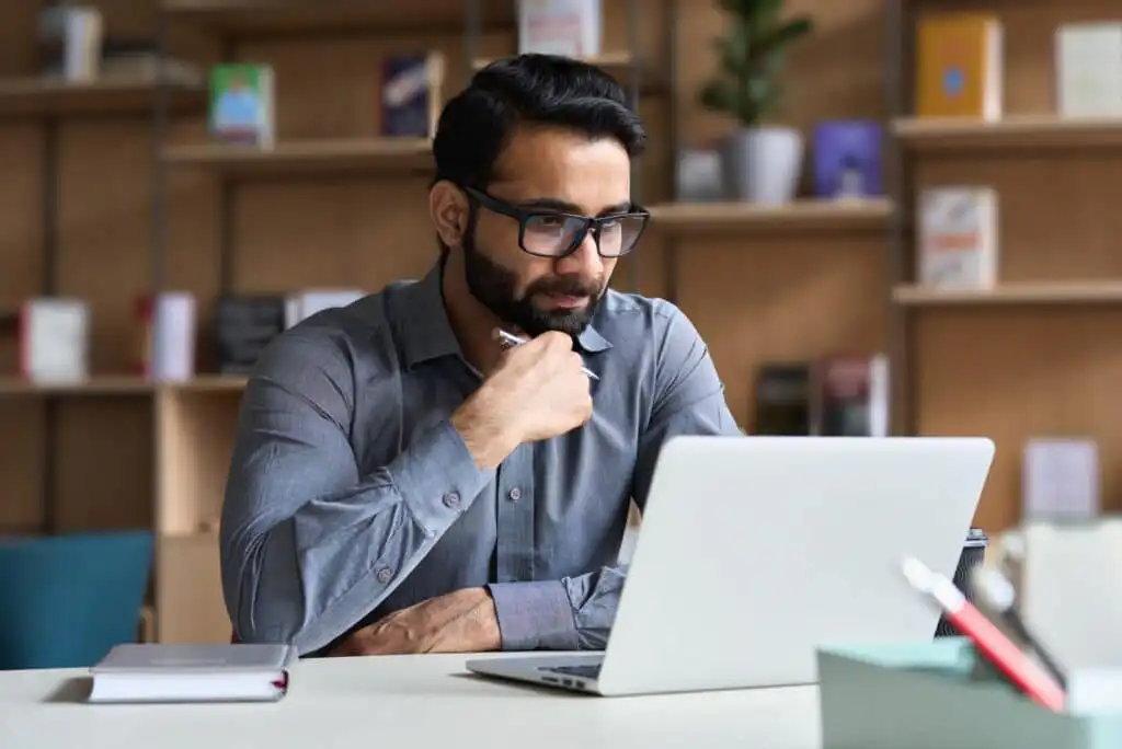 A man with glasses and a beard sits at a desk, looking thoughtfully at a laptop screen. He is wearing a gray shirt, with bookshelves and plants in the background.