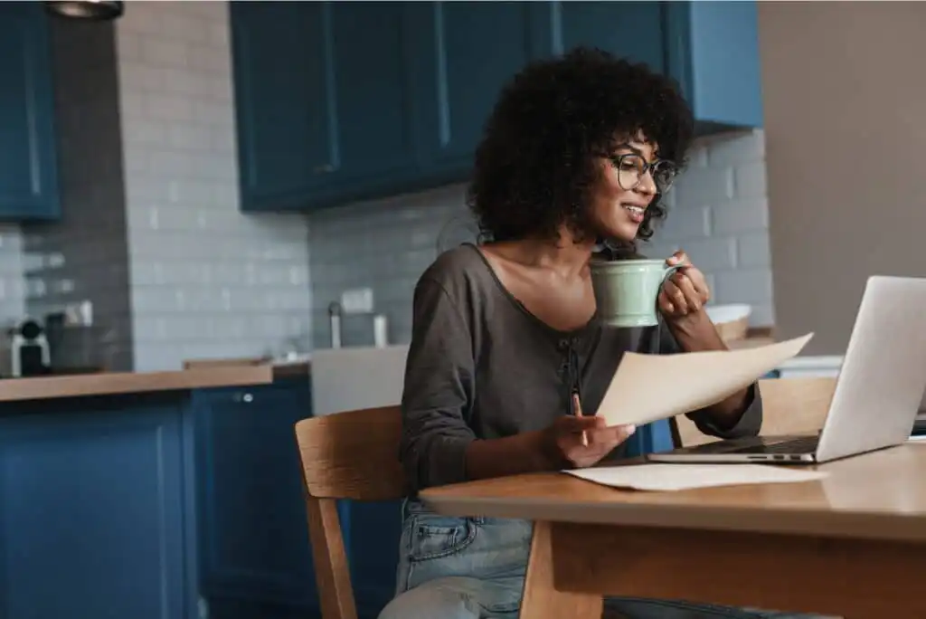 A woman sits at a kitchen table, smiling while holding a mug in one hand and papers in the other, looking at her laptop. The kitchen has blue cabinets and a white tiled backsplash.