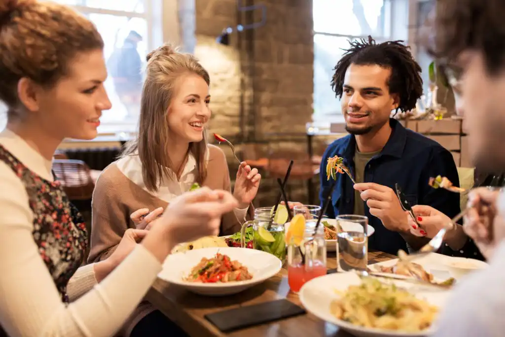 Four young adults sit around a table at a restaurant, smiling and talking while eating pasta dishes. Drinks and plates of food are on the table, and the atmosphere is warm and friendly.