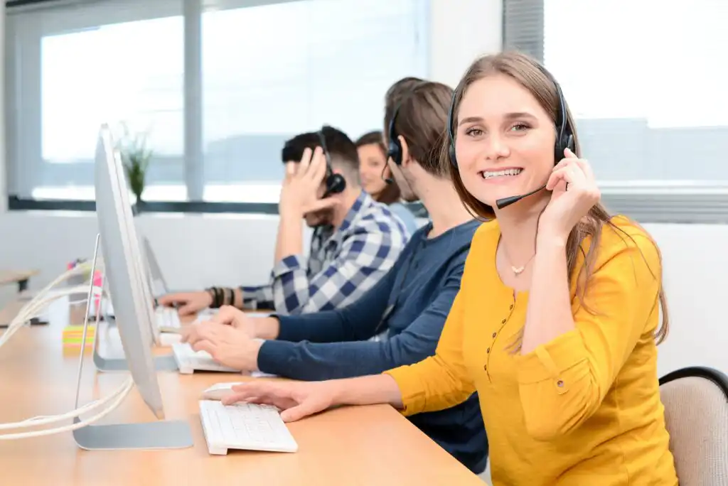 A smiling woman wearing a headset sits at a desk with a computer in an office, alongside several colleagues also wearing headsets and working at their computers.