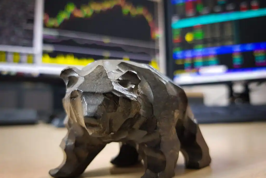 A metal bear figurine sits on a desk with blurry computer monitors displaying colorful stock market charts and data in the background.