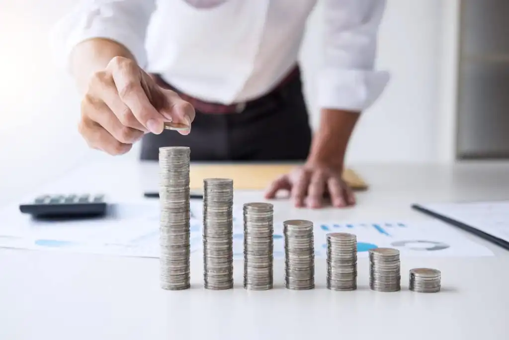 A person stacks coins in ascending order on a desk, with business documents and a calculator in the background, symbolizing financial growth or investment planning.