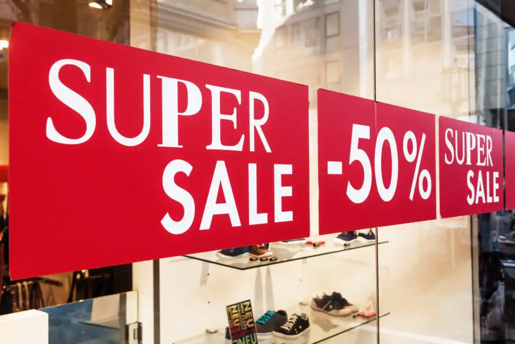 Red signs in a store window display with white text reading “SUPER SALE -50% SUPER SALE,” indicating a significant discount. Reflections of buildings and store items are visible through the glass.