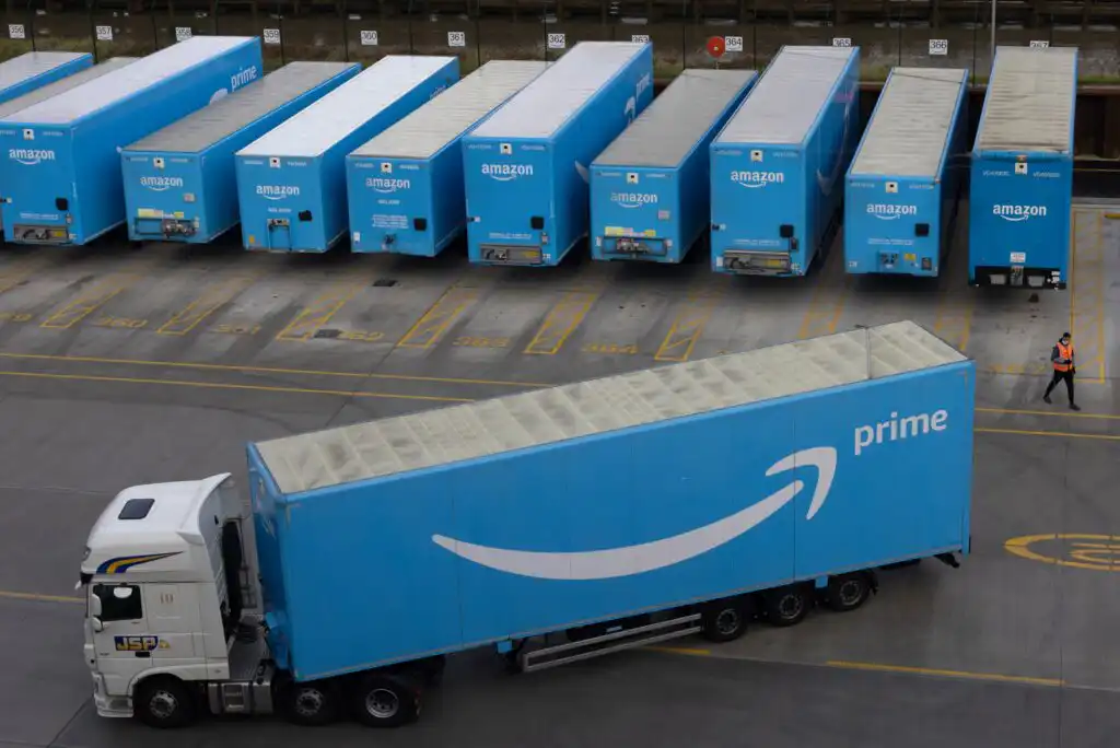 A white truck pulls a blue Amazon Prime trailer in front of several parked blue Amazon Prime trailers in a lot, with a person in an orange vest walking nearby.