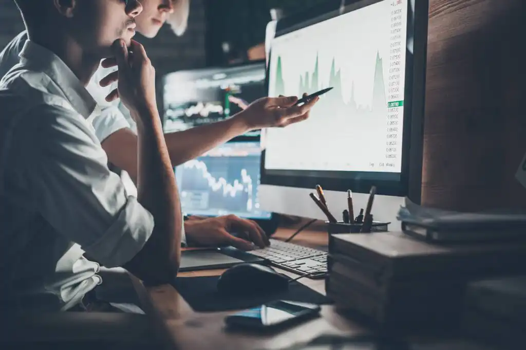 Two people sit at a desk analyzing financial graphs on large computer monitors; one person points at the screen with a pen while the other looks thoughtfully, surrounded by notebooks, pens, and a smartphone.