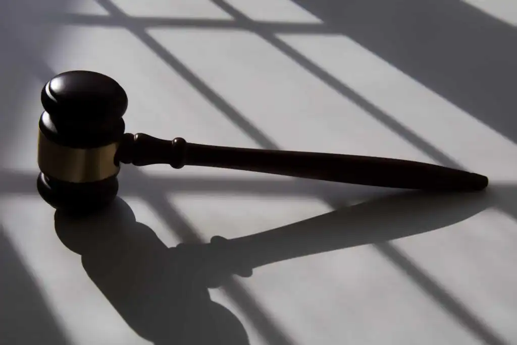 A judge’s gavel lying on a white surface, casting a shadow in the light streaming through a window with bars, creating geometric shadow patterns.