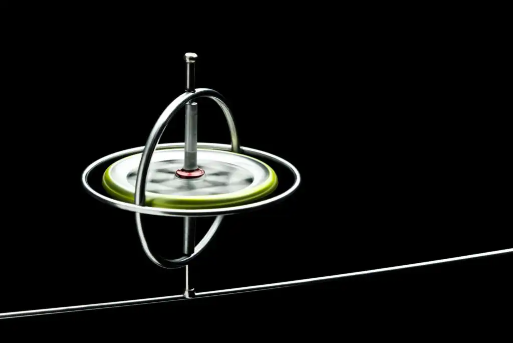 A spinning gyroscope with a green rim and silver frame is balanced on a thin metal wire against a black background.