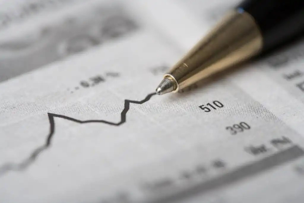 A close-up of a gold pen pointing at a fluctuating line graph on a sheet of paper, possibly representing stock market trends or financial data, with numbers displayed along the side.