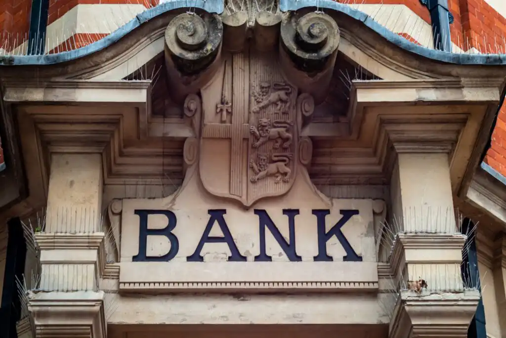 A close-up view of a stone bank building facade featuring the word BANK in large black letters beneath a decorative crest with lion figures and ornate architectural details.