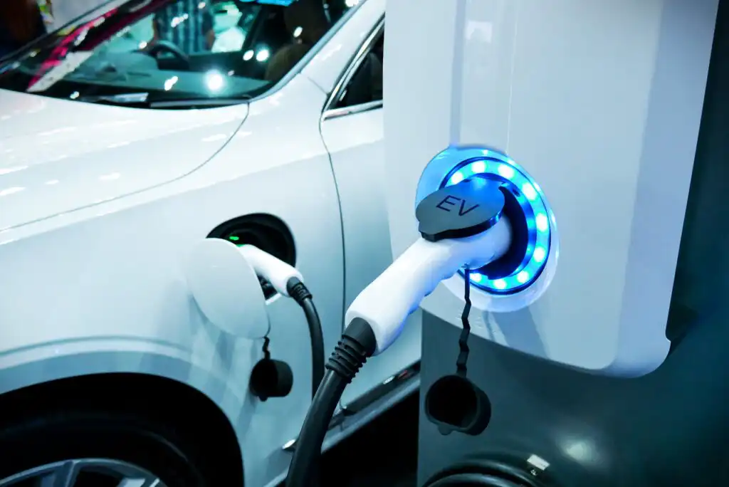 A close-up of an electric car being charged, with a charging cable plugged into the vehicle and a glowing blue ring around the charging port. The white car and charging station are visible.