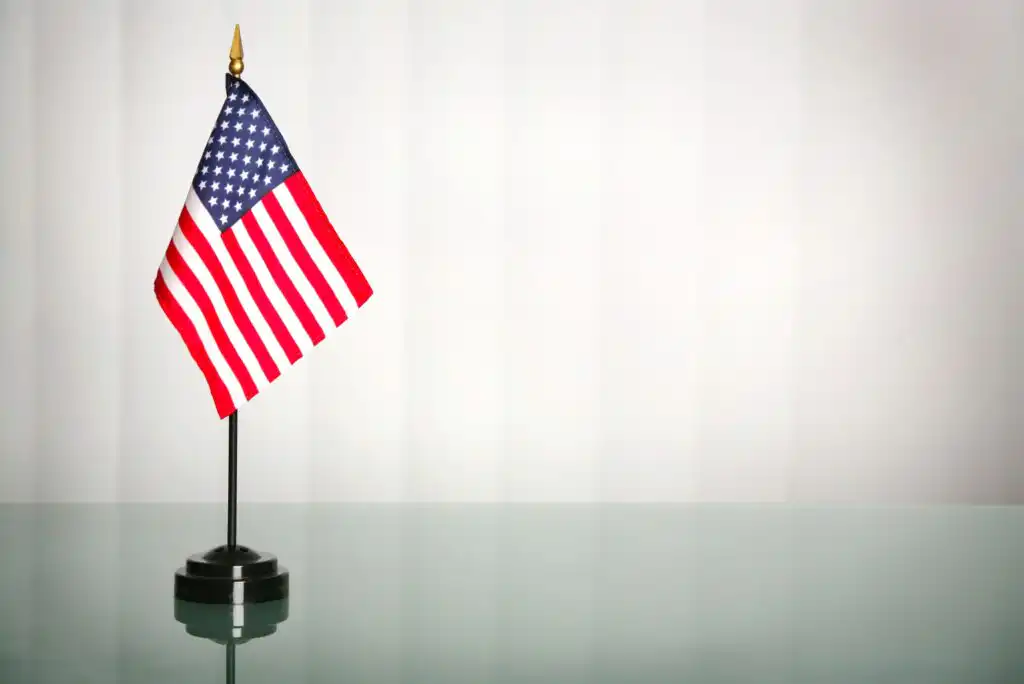 A small United States flag on a black stand sits on a reflective glass surface, with a plain light gray background behind it.