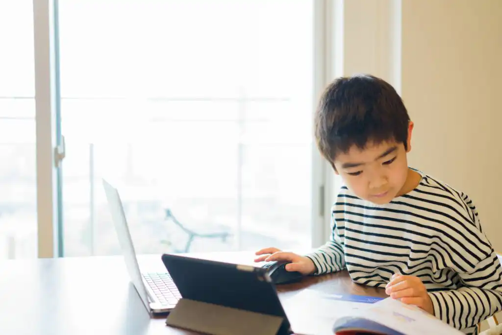 A young boy in a striped shirt sits at a table, looking at a book while using a laptop and tablet. Sunlight streams in through large windows behind him, brightening the room.