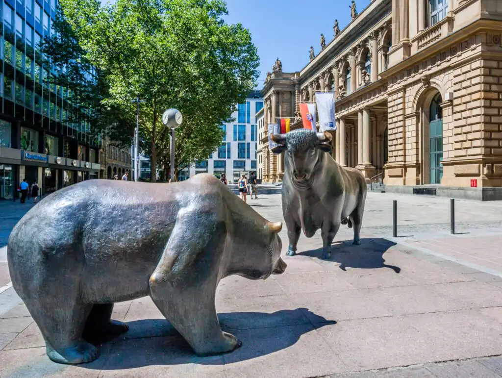 Sculptures of a bear and a bull face each other outdoors in a plaza near the Frankfurt Stock Exchange, surrounded by trees and historic buildings on a sunny day.