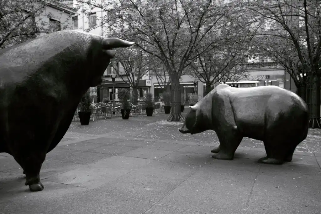 A black and white photo of large bull and bear statues facing each other in an outdoor plaza with trees and buildings in the background.