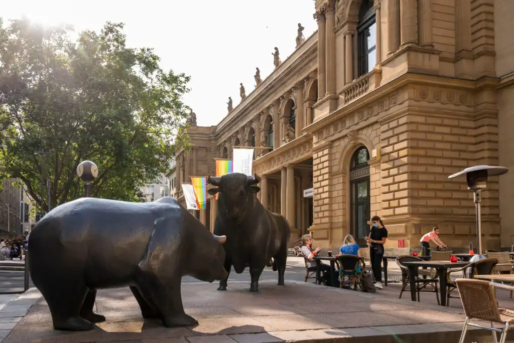 Two large bronze bear and bull statues are displayed outdoors in front of a historic stone building, with people sitting at nearby outdoor tables and trees visible in the background.