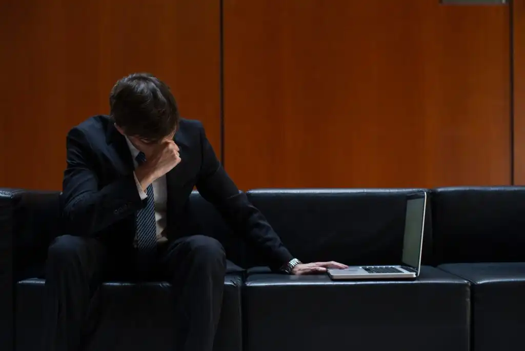 A man in a suit sits on a black couch with his head bowed and hand covering his face, looking stressed. A laptop is open beside him. The background is a plain, orange-brown wall.